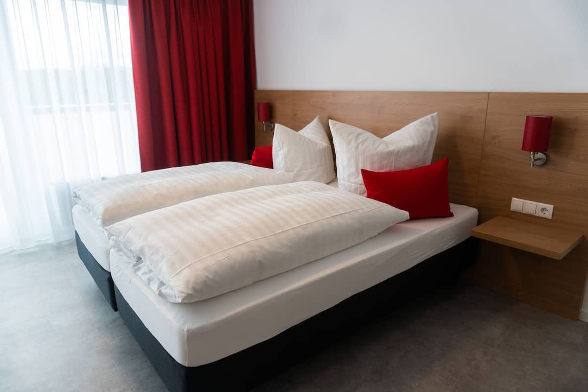 Una cama doble con ropa blanca y almohadas rojas, un cabecero de madera, una mesita de noche con una lámpara y una ventana con cortinas rojas.
