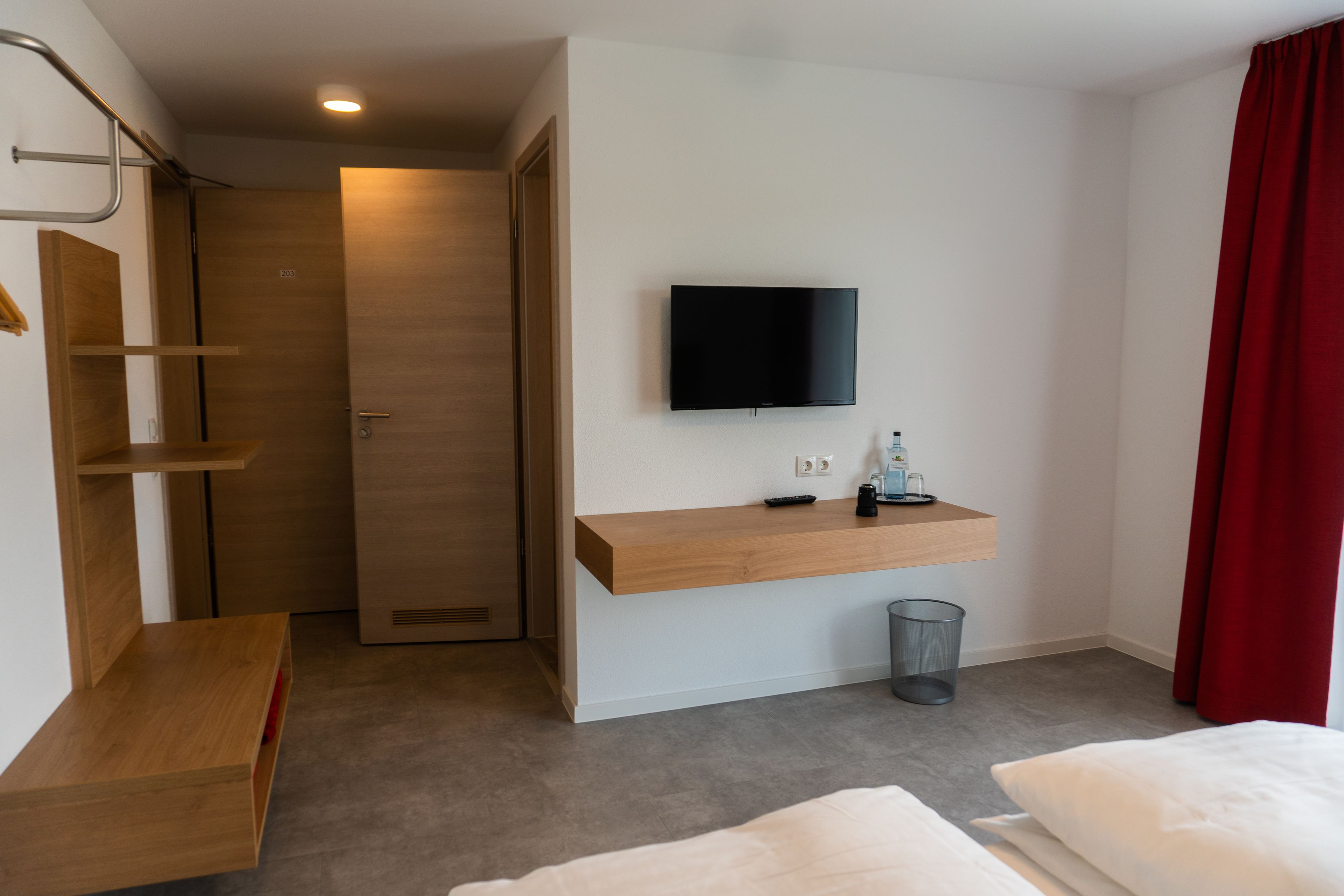 Habitación de hotel con escritorio de madera, televisor montado, pequeño cubo de basura y cortina roja.