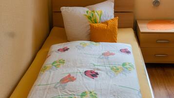 A single bed with a floral bedspread, a white pillow, and a yellow pillow. There is a wooden nightstand with a lamp and a drawer.
