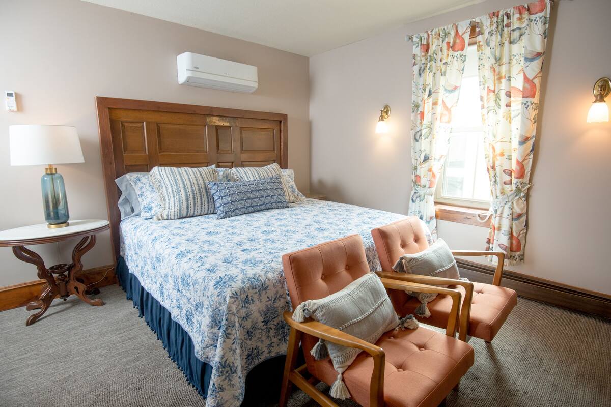 A bedroom with a bed, two chairs, a nightstand, and a lamp.