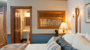 Une chambre à coucher avec un lit, une table de chevet avec une lampe, une tête de lit en bois, une fenêtre ornée de vitraux et une porte donnant sur une autre pièce.