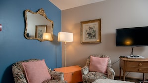 Une chambre d’hôtel avec deux fauteuils à motifs, un petit pouf orange, un lit simple, une télévision, une lampe et un tableau encadré au mur.