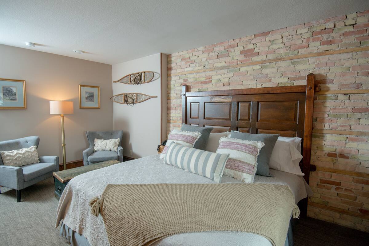 A bedroom with a brick wall, a wooden headboard, a bed with multiple pillows, a side table, and two armchairs.