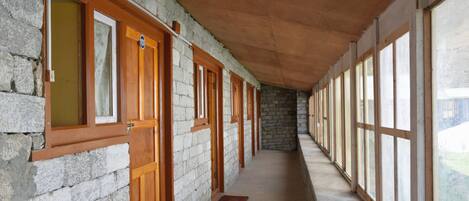 A corridor with stone walls and wooden doors, leading to a room with large windows.