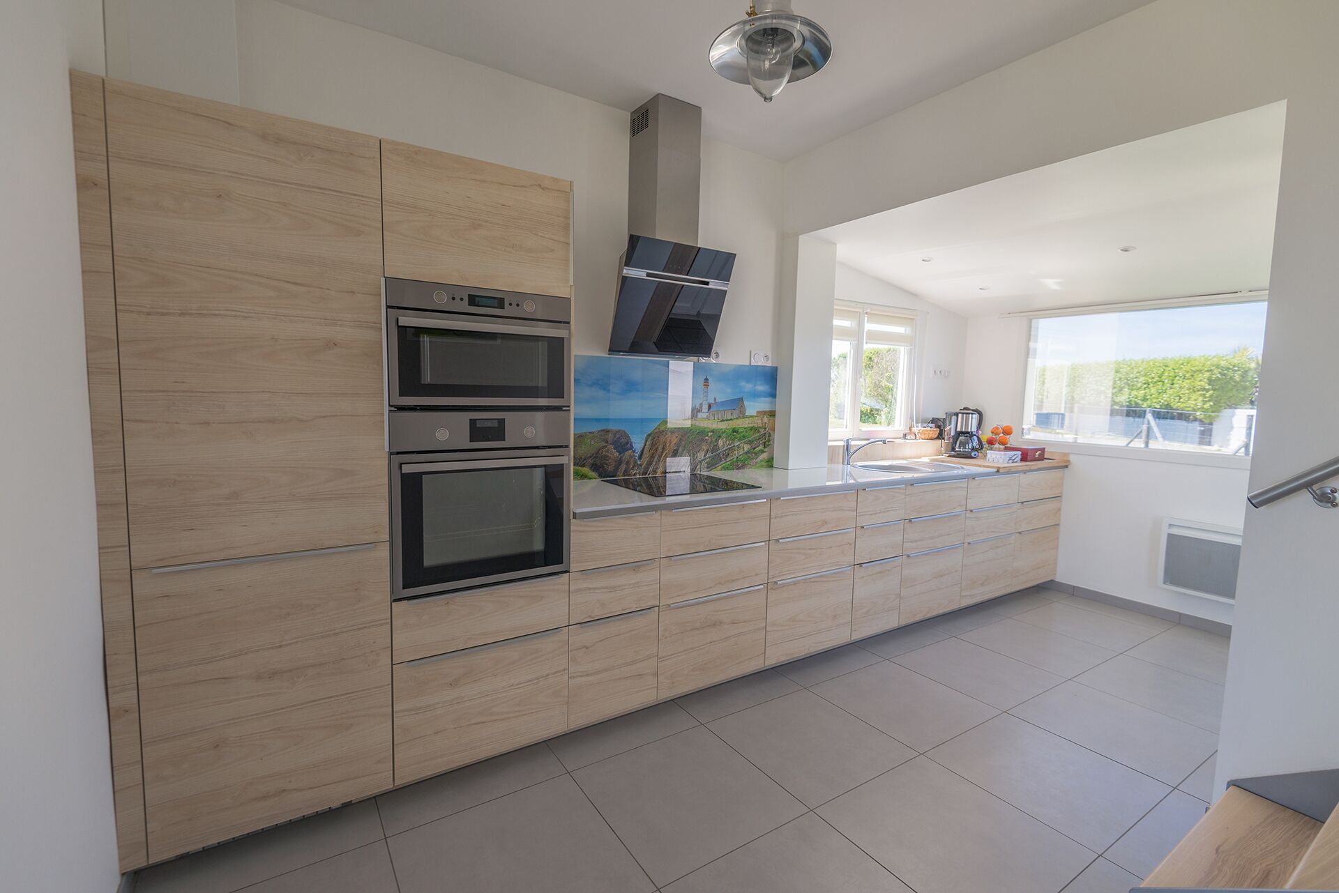 A modern kitchen with wooden cabinets, a double oven, a stainless steel range hood, and a large window offering a view of the outdoors.
