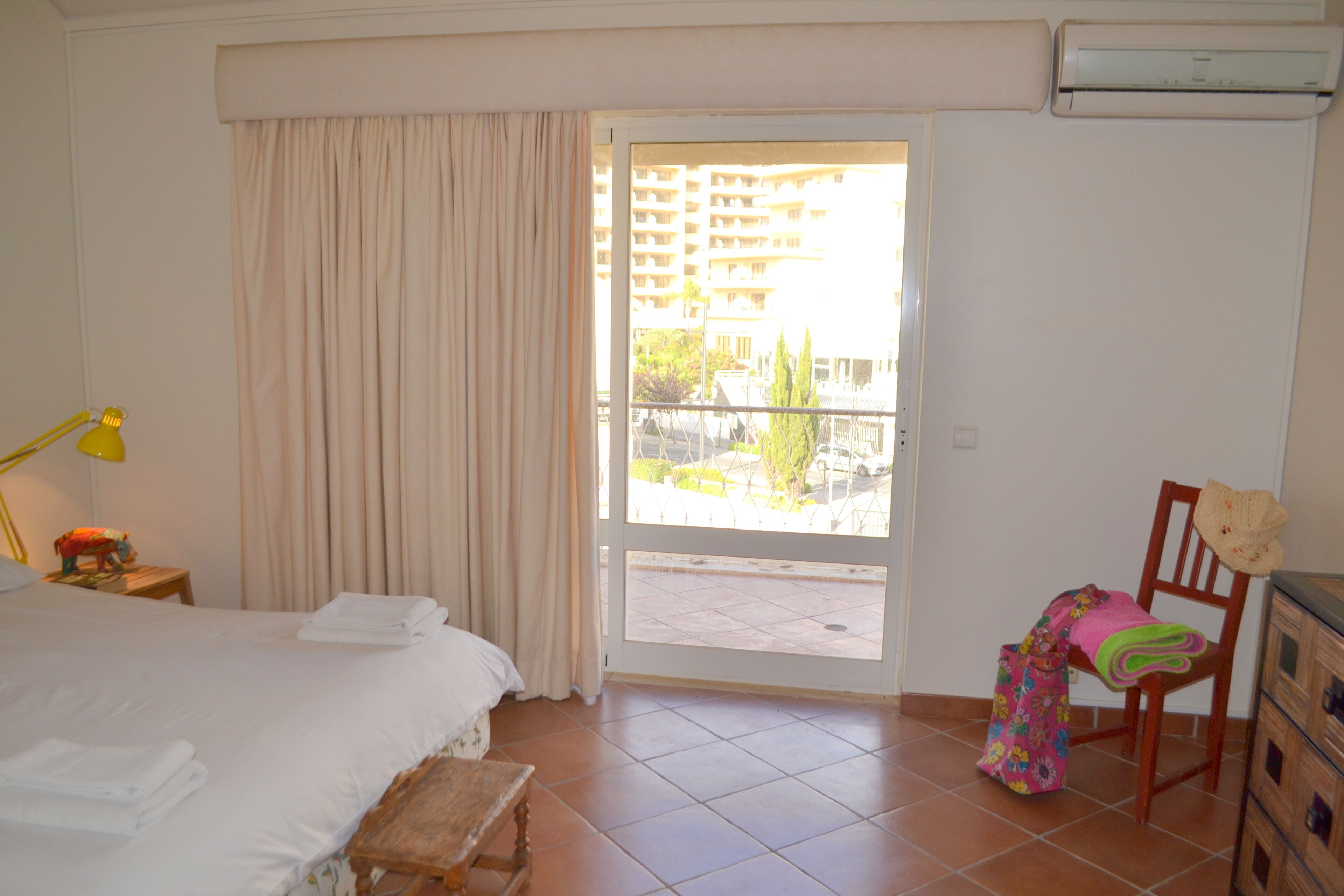 A hotel room with a bed, a wooden nightstand, a chair, a dresser, and a window with a view of a building and trees.