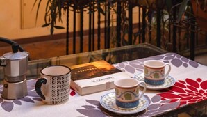 Ein Tisch mit einer Kaffeemaschine, einem Buch und zwei Tassen auf einer gemusterten Tischdecke.