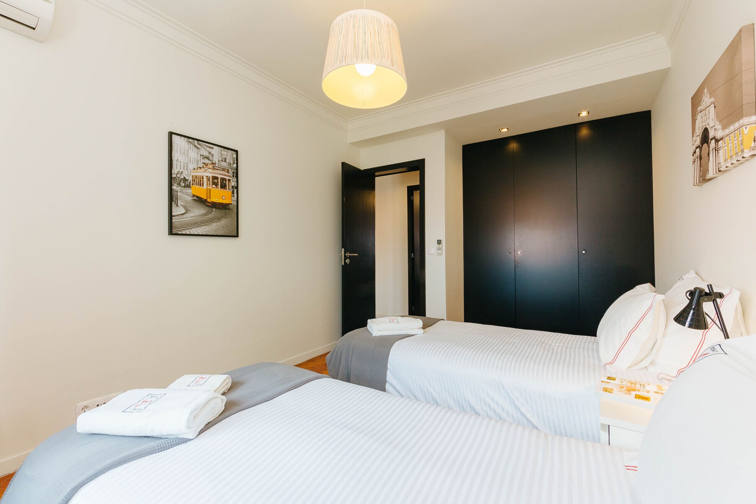 Une chambre d’hôtel avec deux lits, une grande armoire sombre et une image encadrée accrochée au mur, ainsi qu’une lampe suspendue.