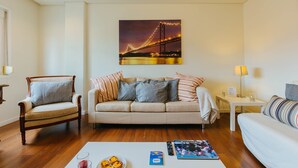 A living room with a sofa, a chair, a coffee table with books and a plate of food, and a painting of a bridge. - RENT4REST LISBON FAMILY & FRIENDS APARTMENT IN ROMA AVENUE (Lisboa ciudad)