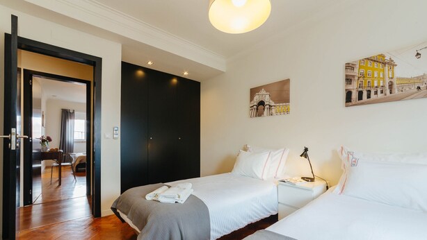 A hotel room with two beds, a wooden floor, a painting of a building, and a door leading to another room. - RENT4REST LISBON FAMILY & FRIENDS APARTMENT IN ROMA AVENUE (Lisboa ciudad)