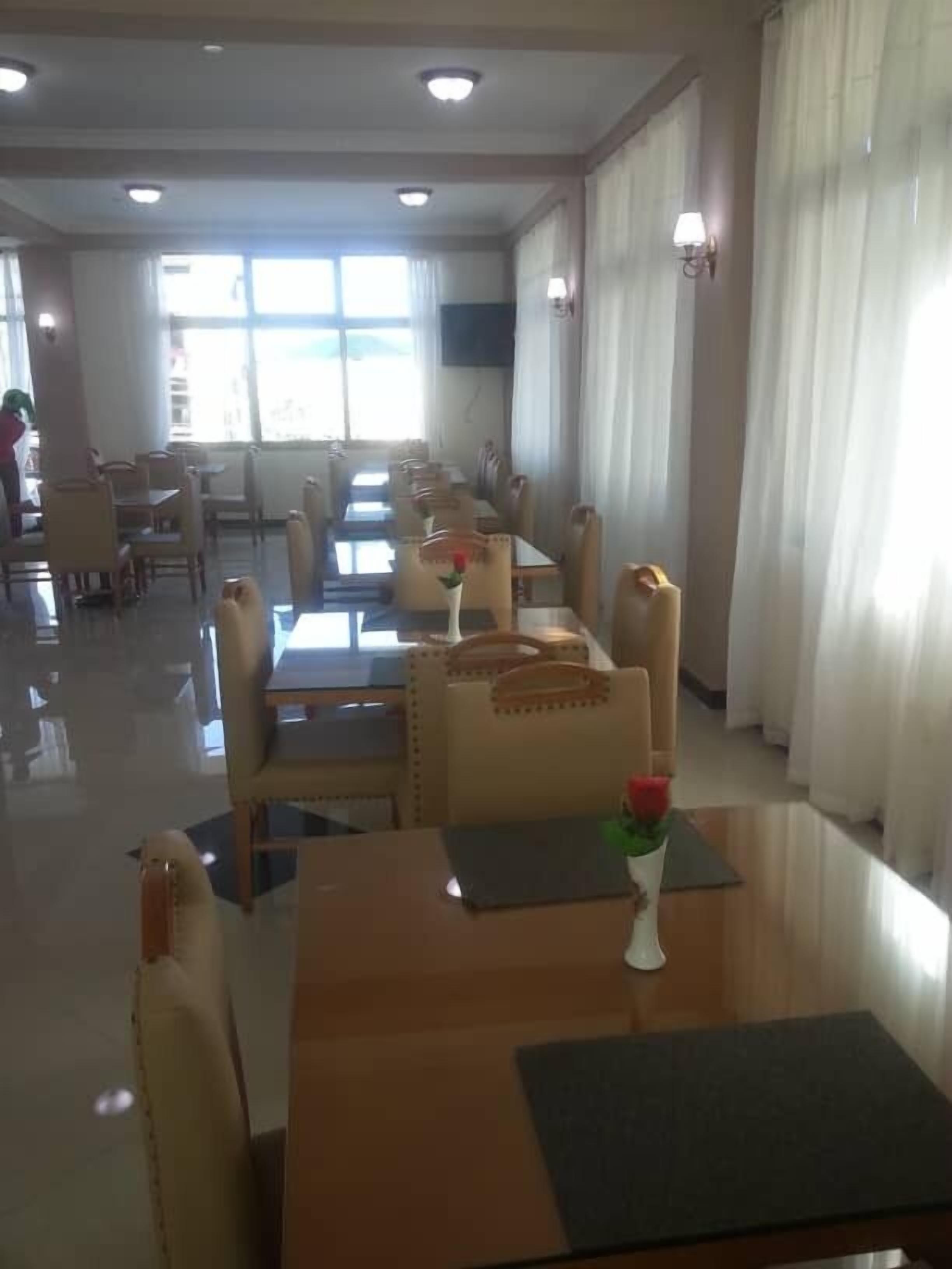 A hotel dining area with tables and chairs, a large window with curtains, and a wall-mounted TV.