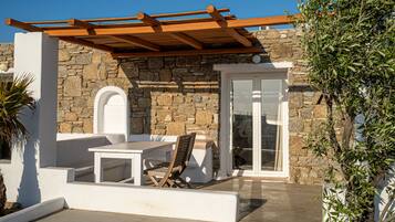 Un edificio de piedra con una pérgola de madera, una mesa blanca y una silla afuera.