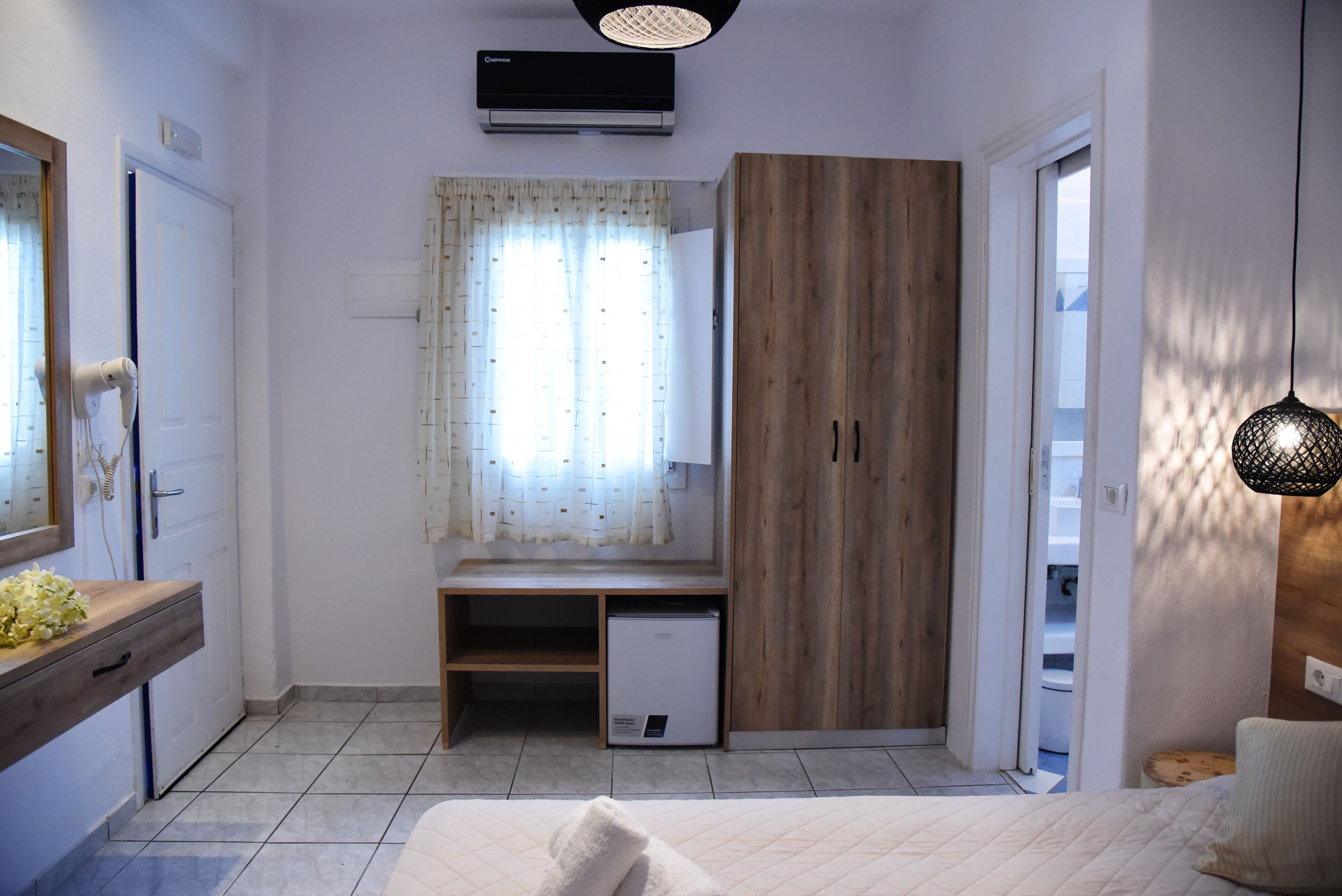 Ein Schlafzimmer mit einem Bett, einem Holzschrank, einem Schreibtisch mit Stuhl, einem kleinen Kühlschrank und einem Fenster mit Vorhängen.