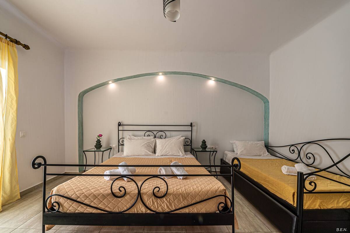 A double bed with a curved headboard, two bedside tables, a small potted plant, and a yellow bedspread.