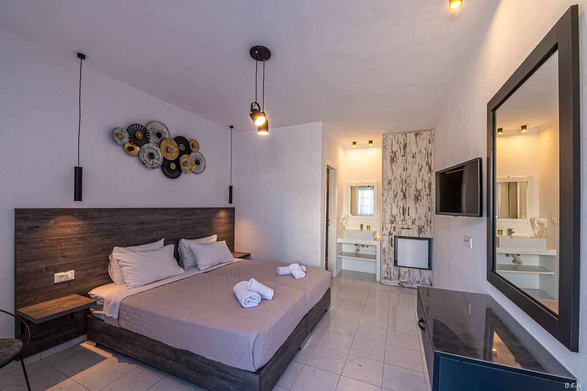 A modern bedroom with a large bed, bedside tables, a TV, a mirror, and a bathroom with a sink and mirror.