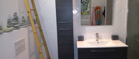 A bathroom with a white sink, dark cabinet, and a mirror. There is a bamboo ladder leaning against the wall and a small shelf with toiletries.