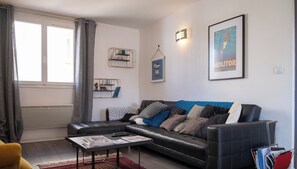 Uma sala de estar moderna com um sofá de couro preto, uma mesa de centro e um tapete vermelho. Há estantes com objetos decorativos e um pôster emoldurado na parede.