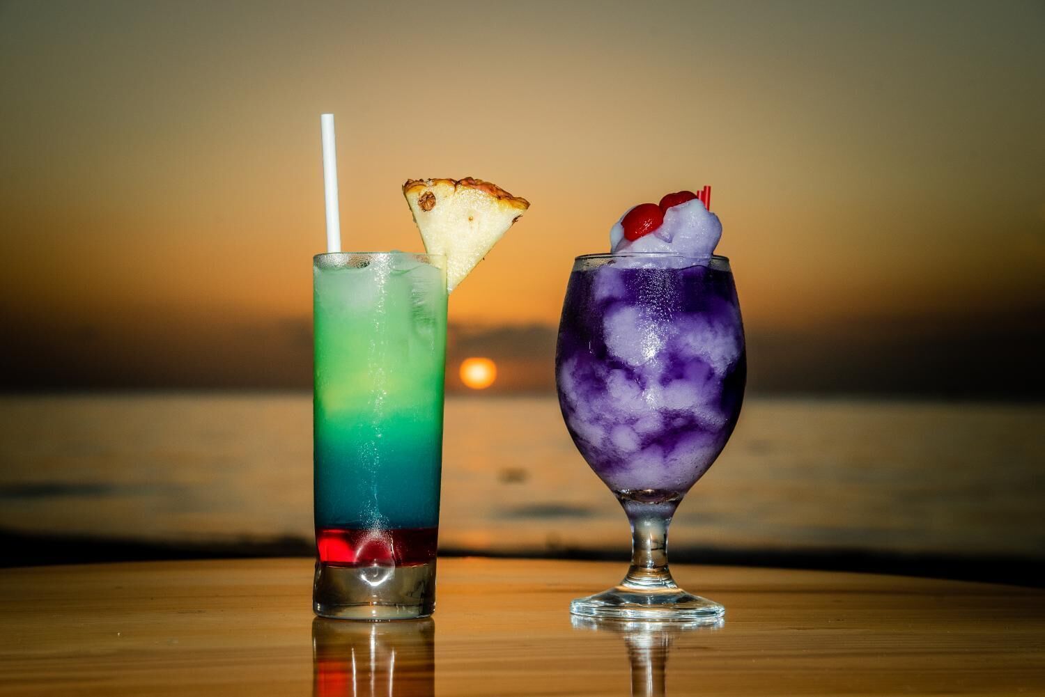 Two cocktails on a wooden table with a sunset in the background.