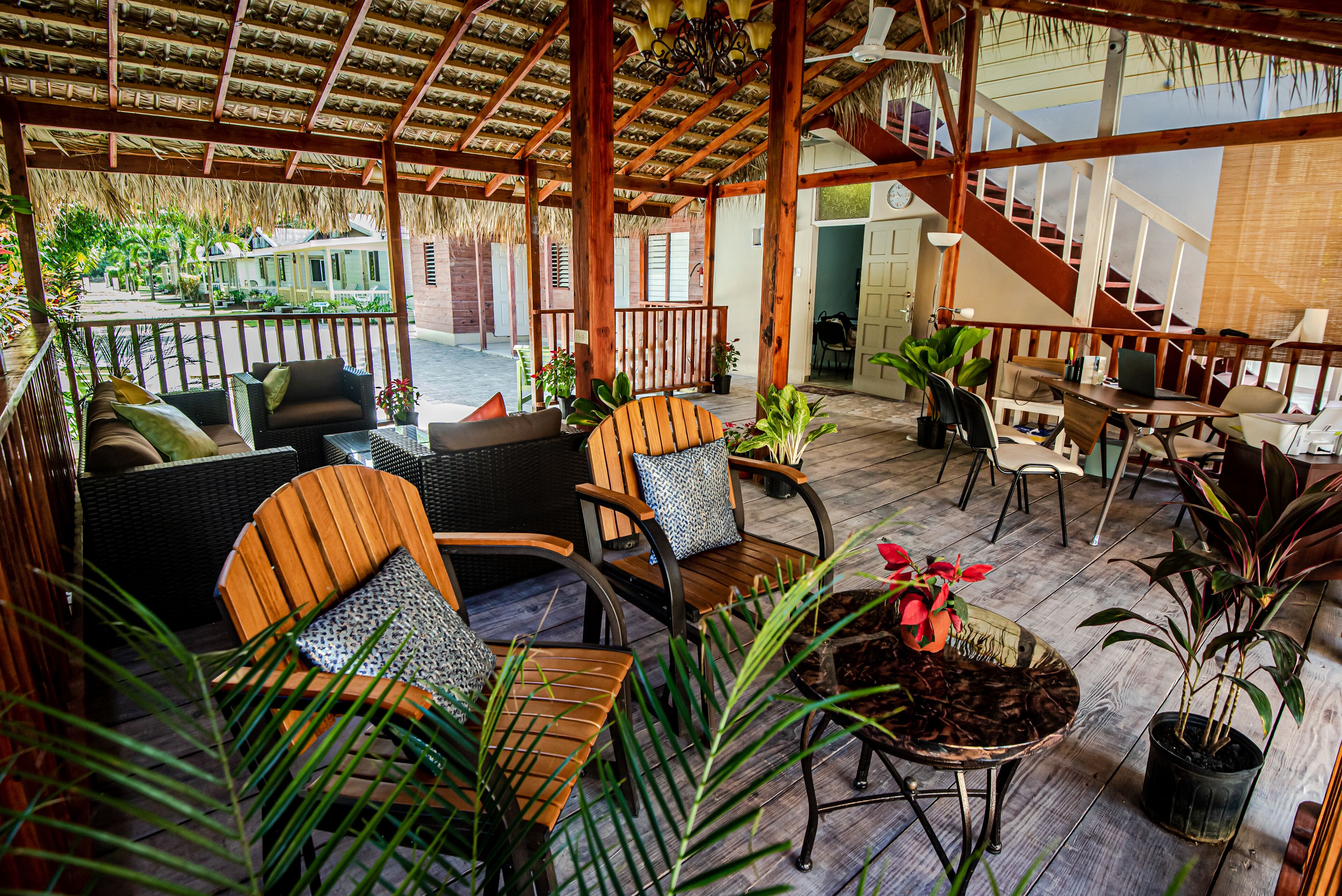 A covered outdoor seating area with wooden furniture, potted plants, and a staircase leading to an upper level.