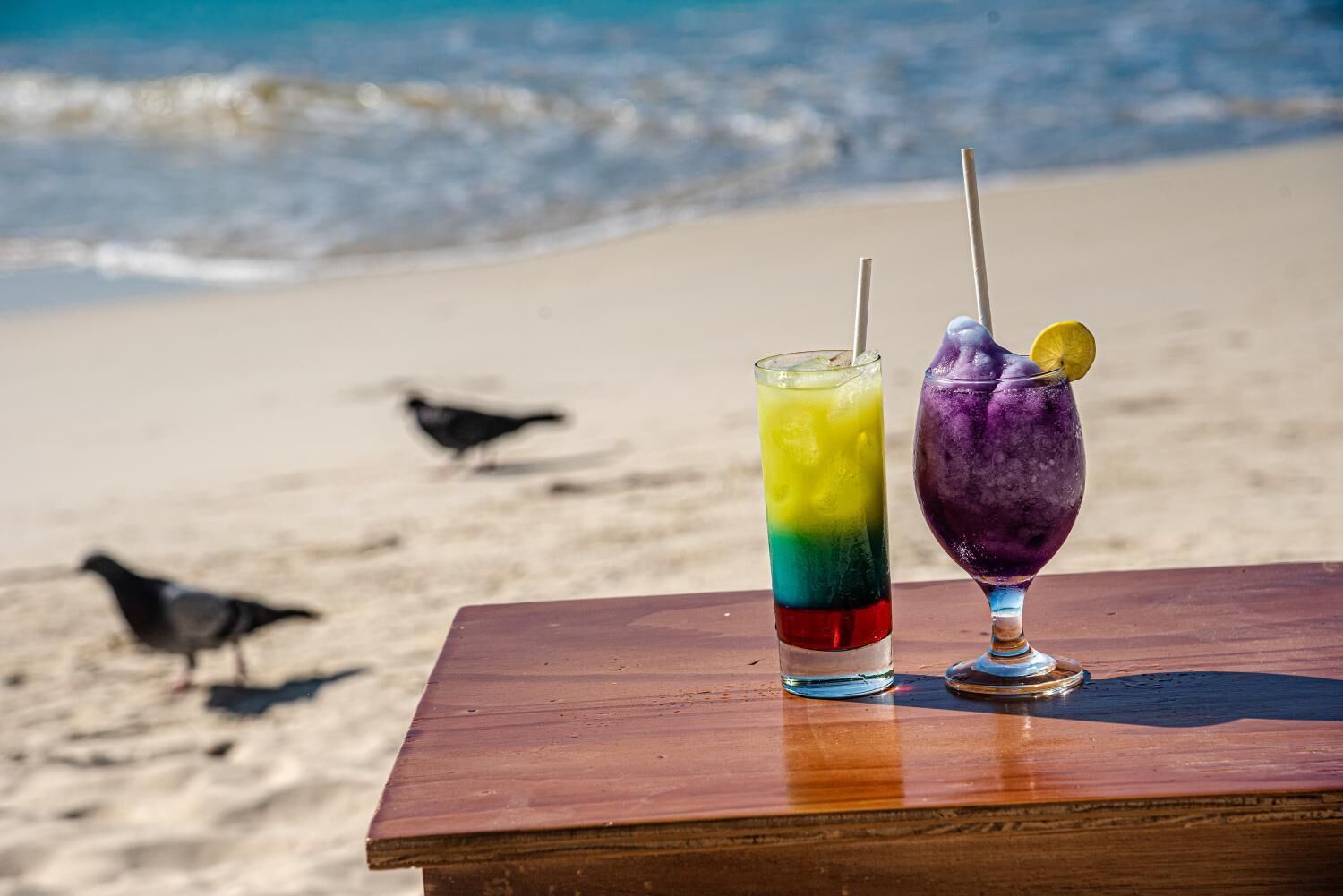 Dos bebidas coloridas sobre una mesa de madera, con una playa y pájaros al fondo.