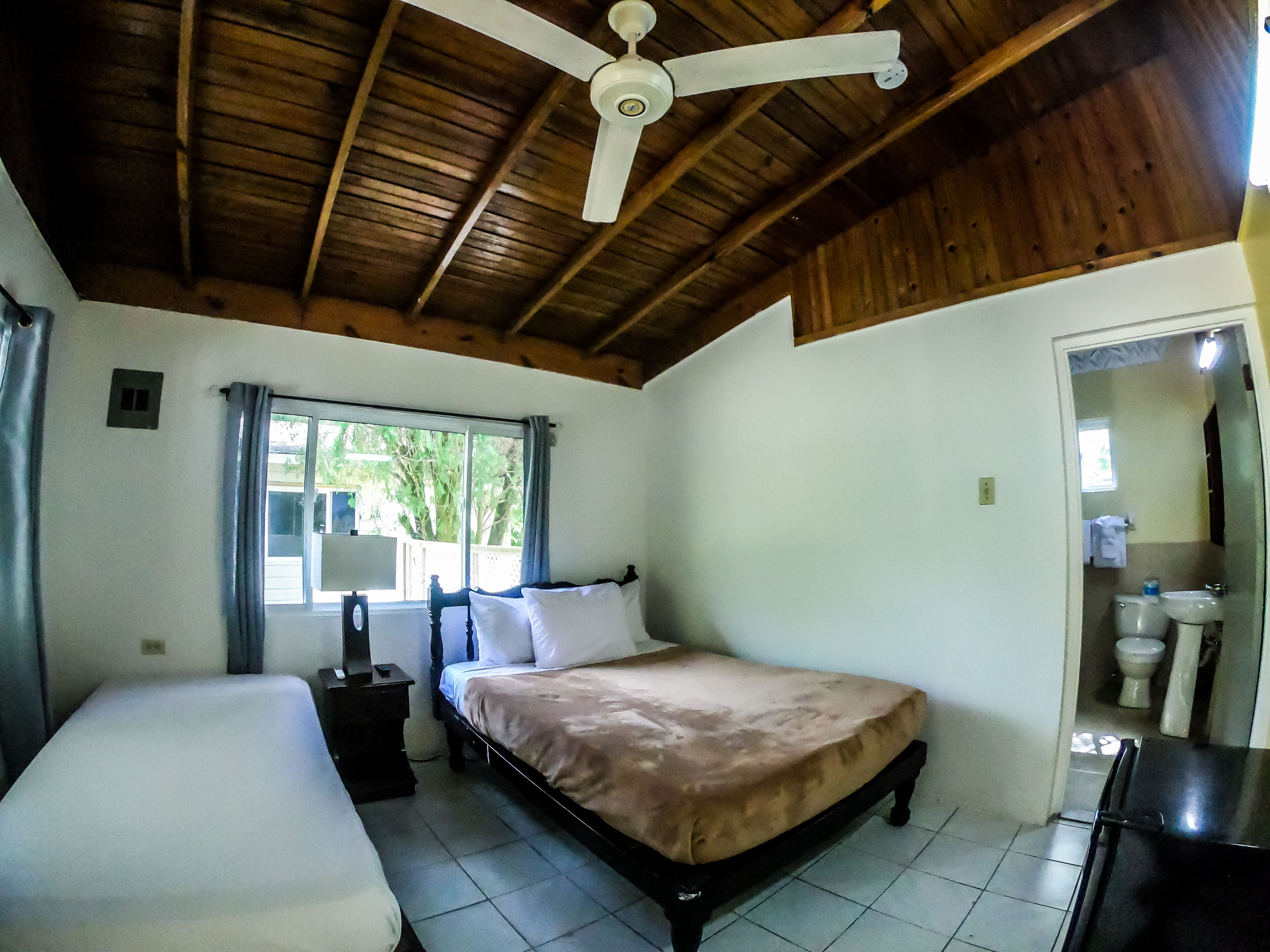 A bedroom with a bed, a bedside table, a ceiling fan, a window with curtains, and a bathroom visible through an open door.