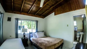 A bedroom with a bed, a bedside table, a ceiling fan, a window with curtains, and a bathroom visible through an open door.