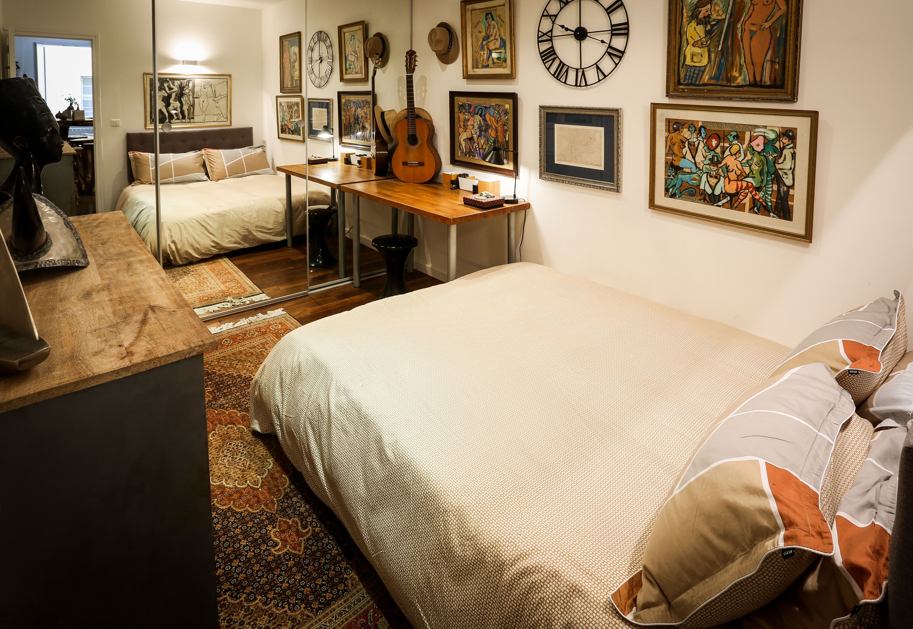 Une chambre à coucher avec un lit, un bureau et une guitare. Plusieurs horloges et des œuvres d’art encadrées ornent le mur.