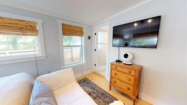 A bedroom with a bed, a TV mounted on the wall, a wooden dresser, and a window with a view of greenery.
