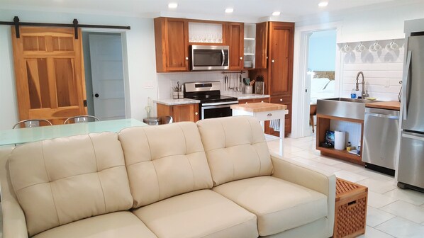 Una cocina moderna con armarios de madera, un refrigerador de acero inoxidable y un comedor con mesa de vidrio y sofá color beige.