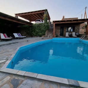 A rectangular swimming pool with a stone border, surrounded by lounge chairs and a covered patio area.