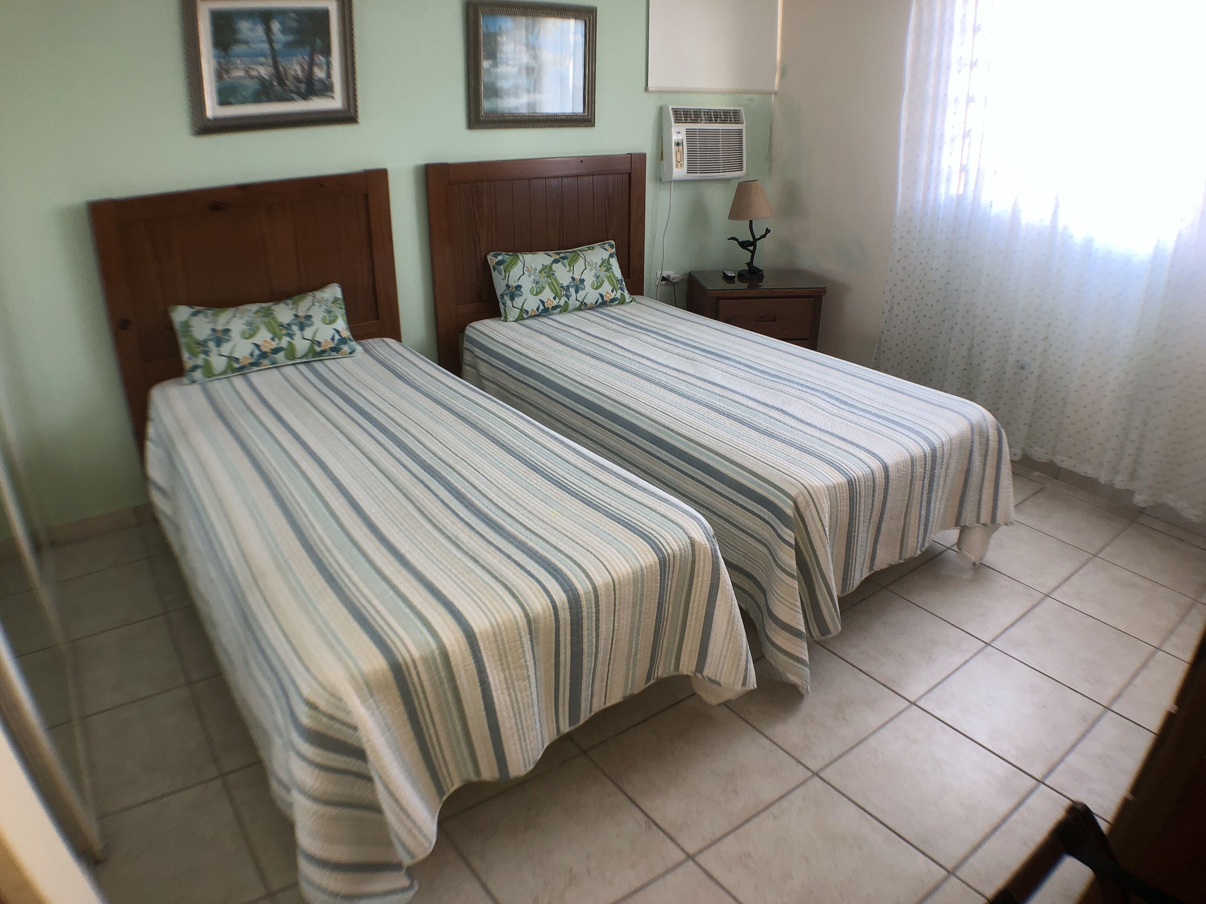 A room with two beds, each with striped bedding and floral pillows. There is a nightstand with a lamp and an air conditioner on the wall.