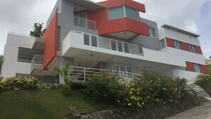 A modern multi-story building with balconies, a stone retaining wall, and yellow flowering plants. - OCEAN VIEW "GATED" AND ACROSS THE WYNDHAM HOTEL!!! (RIO GRANDE RIO MAR)