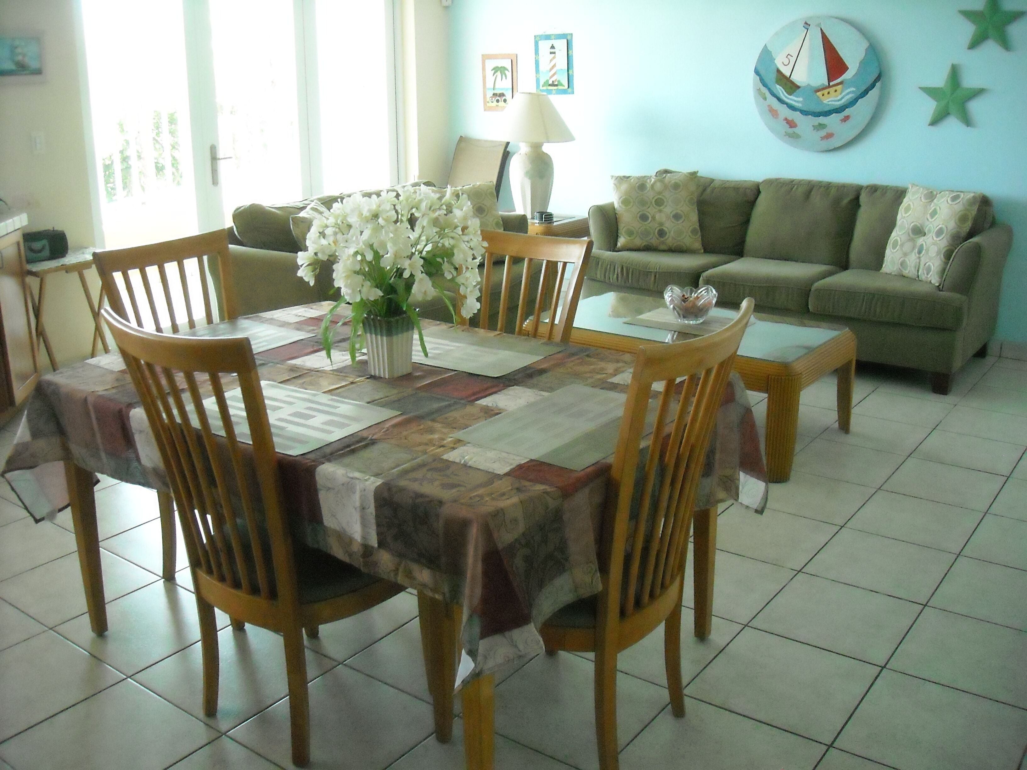 Un coin repas avec une table dressée pour quatre personnes, entourée de chaises en bois, et un salon avec un canapé et des œuvres d’art décoratives accrochées au mur.