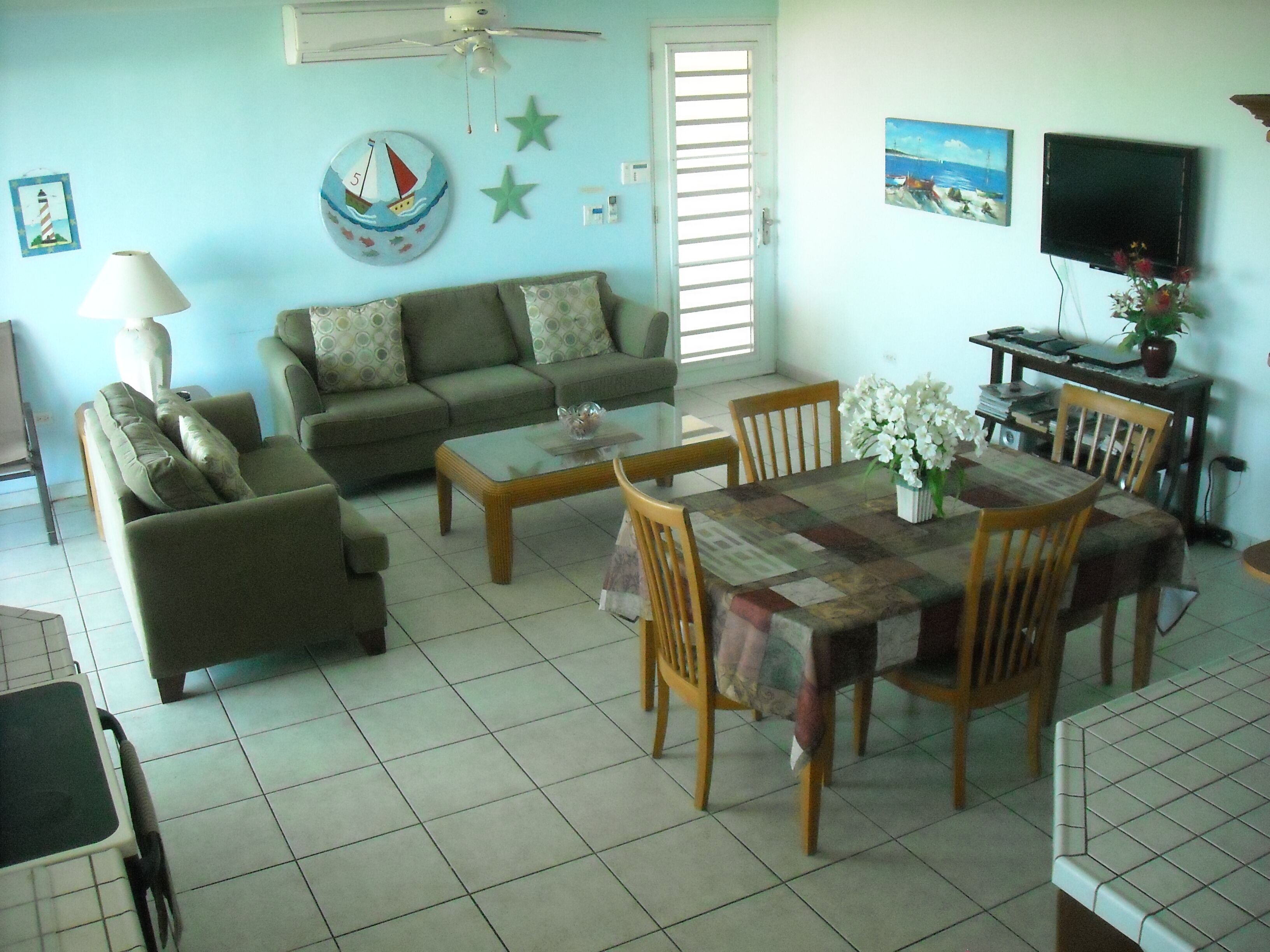 A living room with a sofa, a coffee table, a dining table with chairs, a TV, and a ceiling fan.