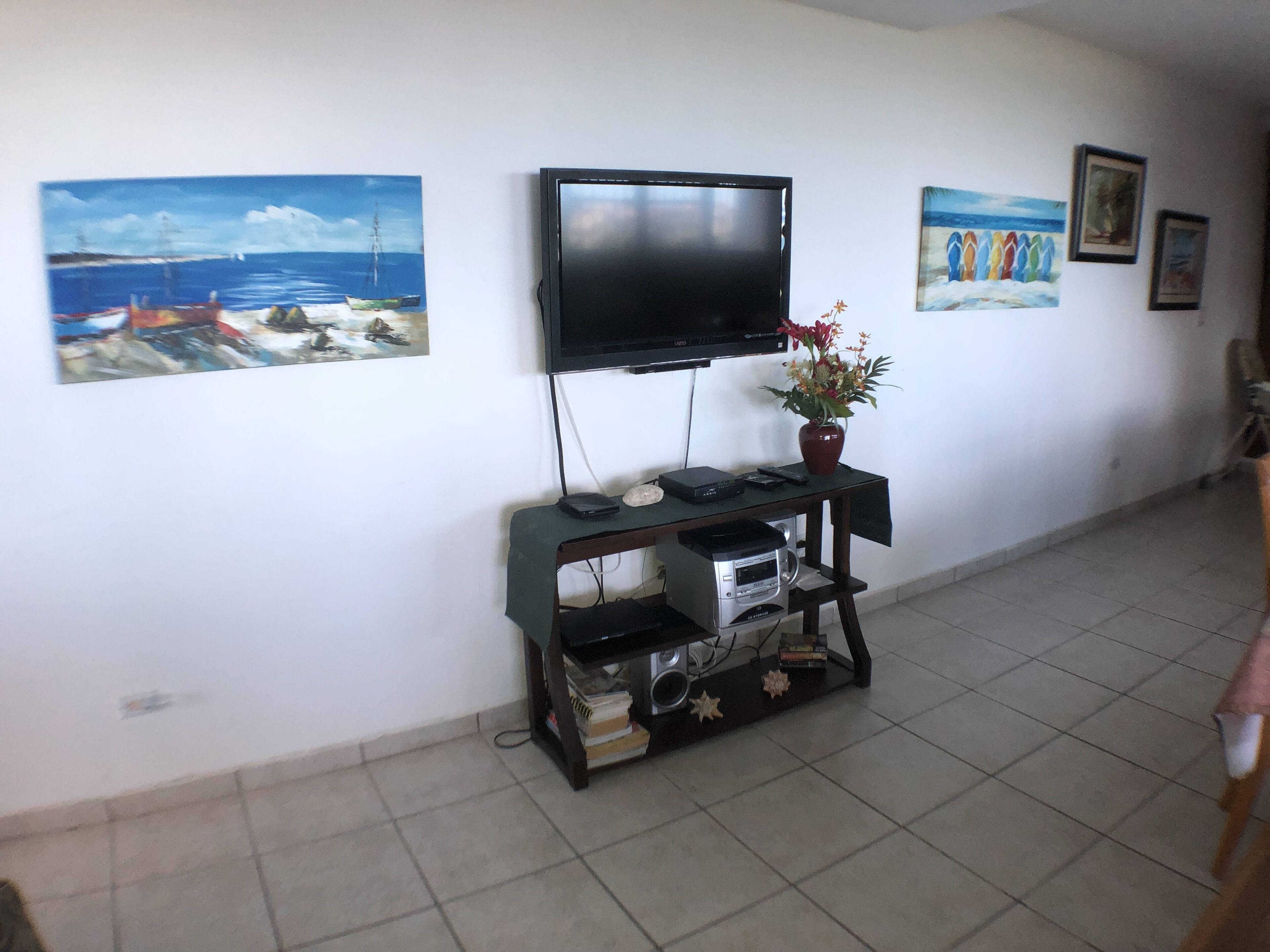 A living room with a flat-screen TV mounted on the wall, a small table with a vase of flowers, and artwork on the walls.