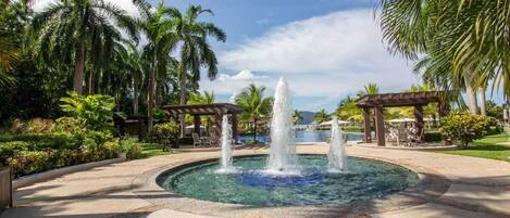 A circular pool with a central fountain, surrounded by palm trees and a gazebo.