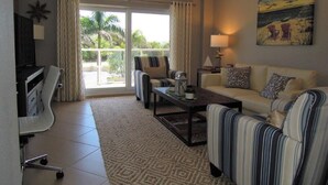 A living room with a sofa, armchairs, a coffee table, and a TV. There is a balcony with curtains and a painting on the wall. - Beachcomber unit 201 (Longboat Key)