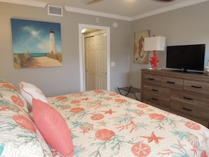 Une chambre à coucher comprenant un lit, un téléviseur, une commode, un tableau représentant un phare et une porte donnant sur une autre pièce.