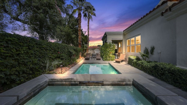 Una piscina con zona de patio, rodeada de exuberante vegetación y una casa de fachada blanca y tejados de tejas marrones.