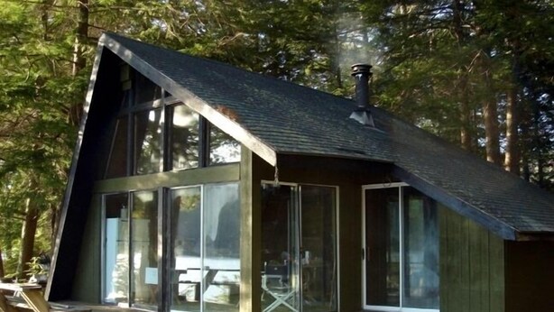A modern cabin with large glass windows, a wooden deck, and a sloped roof.