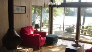 A cozy room with a wood-burning stove, a red armchair, a blue ottoman, and a view of a lake through large windows.