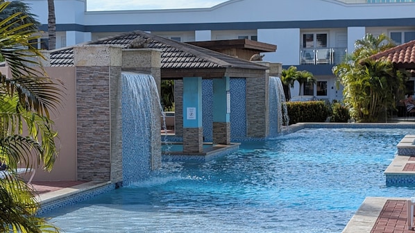 Una piscina con caída de agua, rodeada de palmeras y un edificio con techo de tejas.