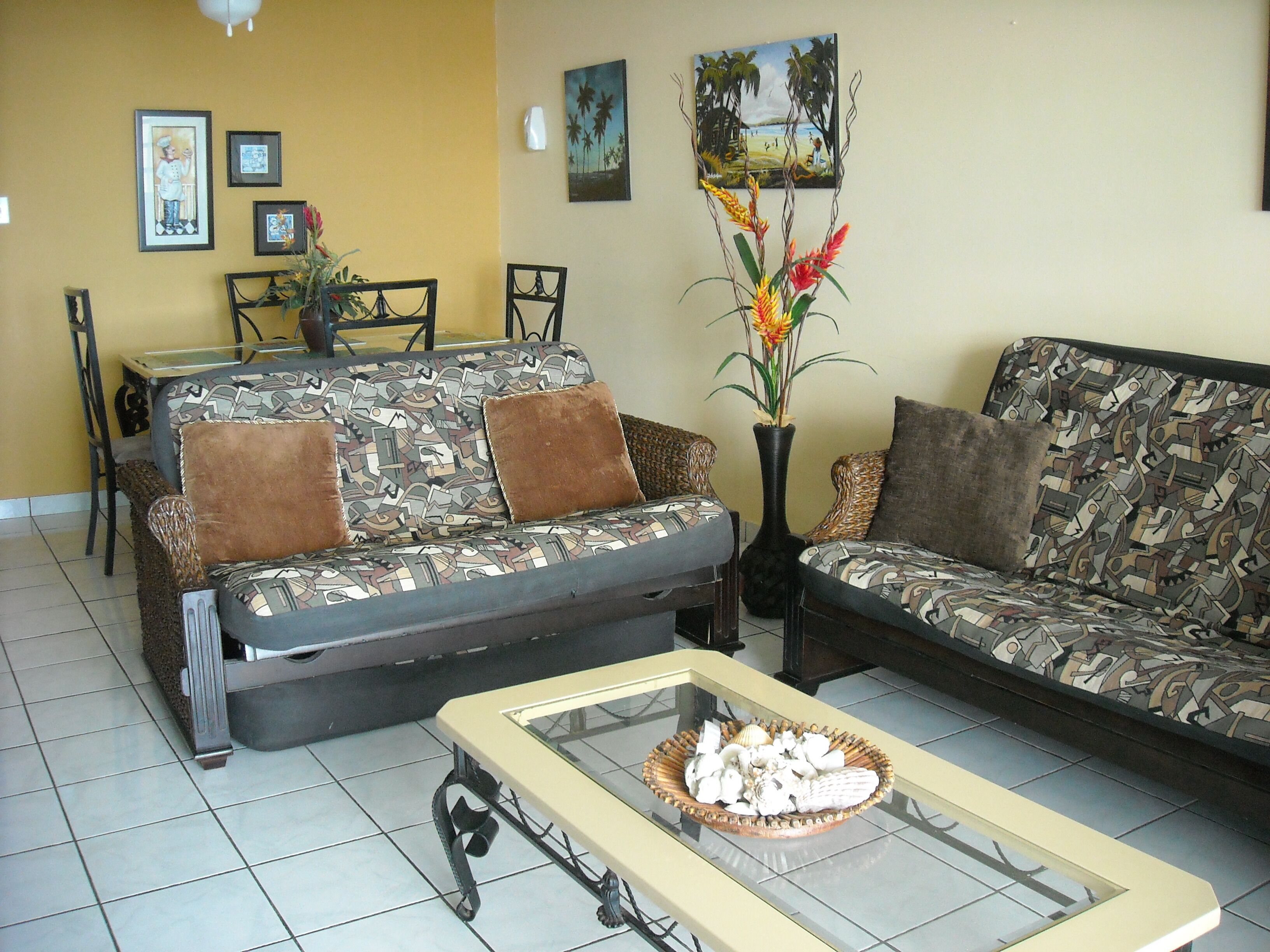 A living room with two patterned sofas, a glass coffee table with decorative items, a dining area with chairs and a table, and wall art including a beach scene.