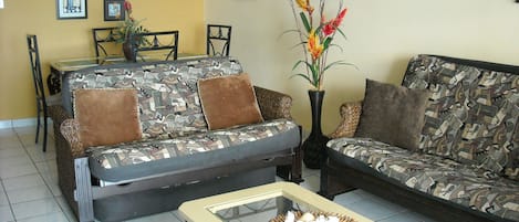 A living room with two patterned sofas, a glass coffee table with decorative items, a dining area with chairs and a table, and wall art including a beach scene.