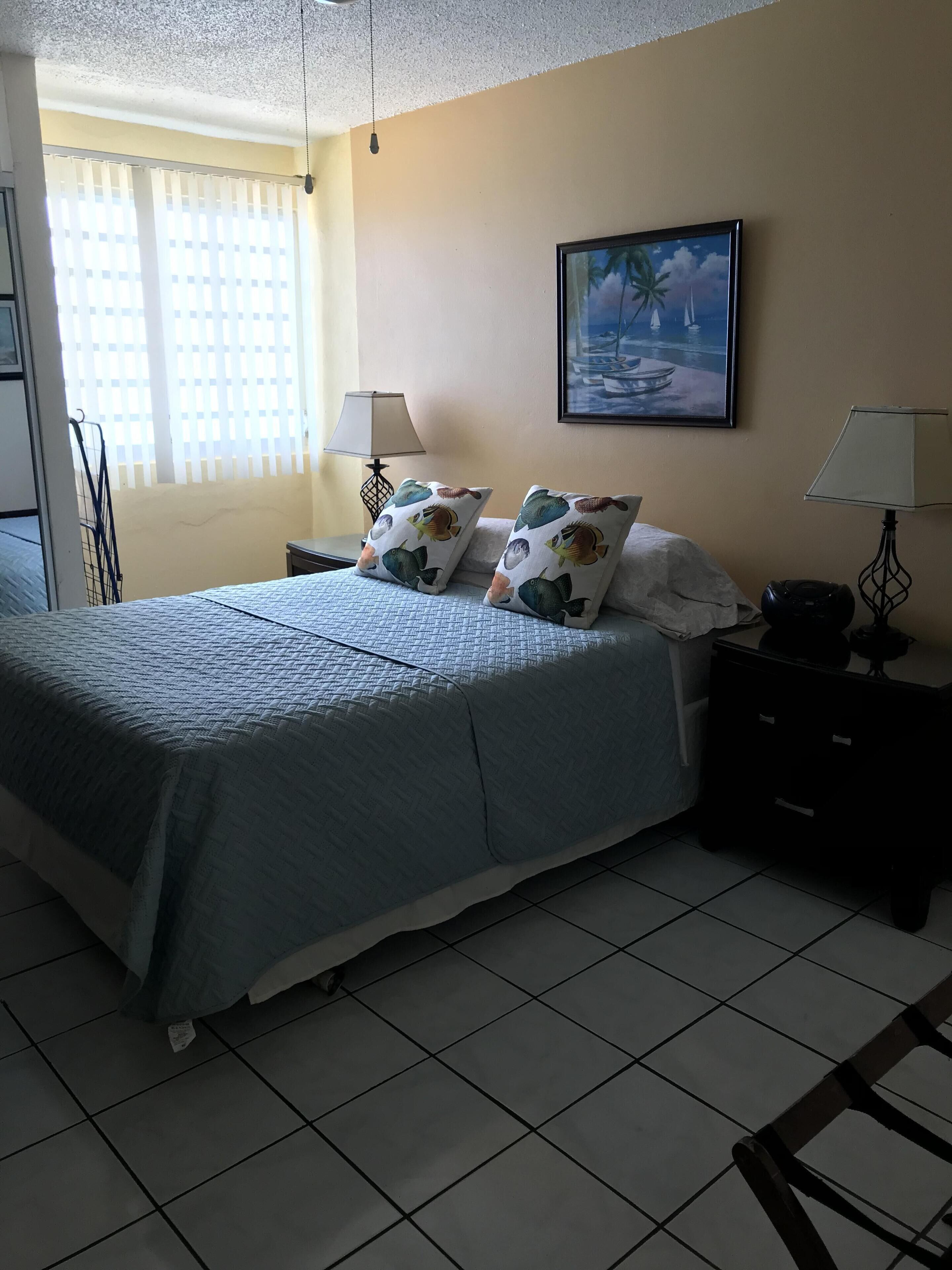 Une chambre à coucher bien rangée, avec un lit, deux tables de chevet, une lampe, un tableau et une fenêtre avec des stores.