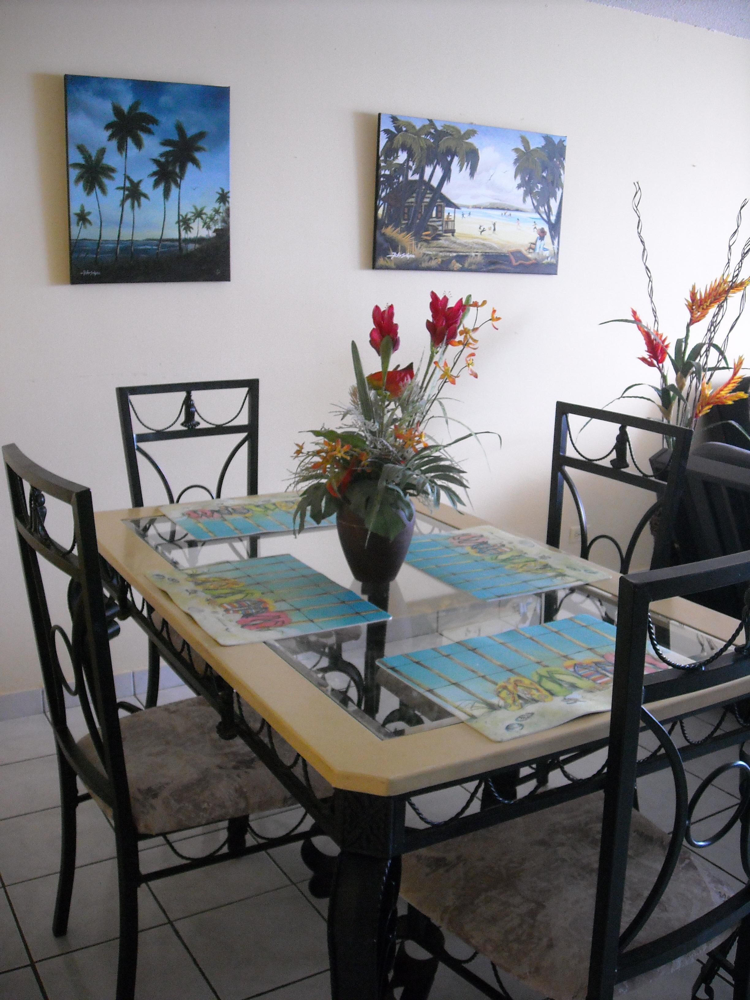 Uma mesa de jantar com um arranjo floral no centro, rodeada por cadeiras, e duas pinturas na parede ao fundo.
