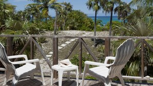 Une terrasse en bois, avec des chaises en plastique blanches et une petite table, donnant sur une plage bordée de palmiers et sur l’océan.