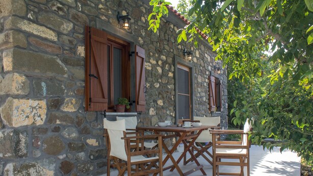A stone building with wooden shutters, a table with chairs, and a potted plant. - 'Perivoli' Stone House (Eresos Lesbos)
