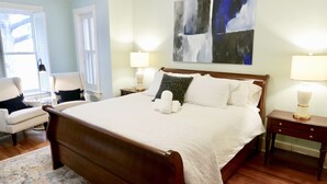 A neatly arranged bedroom with a large bed, two bedside tables with lamps, a chair, and a window with shutters.