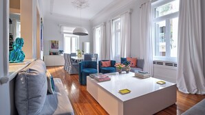A spacious living room with a large white coffee table, blue and grey sofas, and a dining area with a white table and chairs. - Acropolis Whitehouse Mansion in Plaka by JJ Hospitality (Athina)